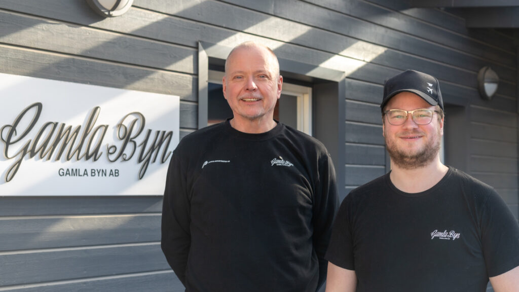 Två män i svarta kläder mot en mörkgrå träfasad.