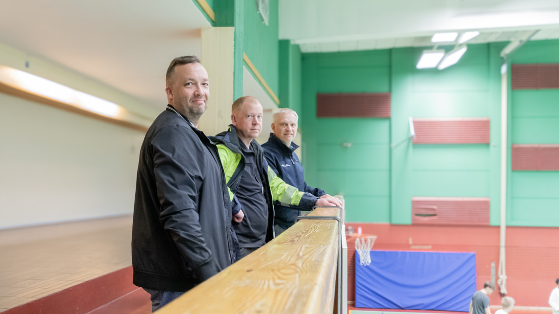 Tre män står på en läktare i en idrottshall.