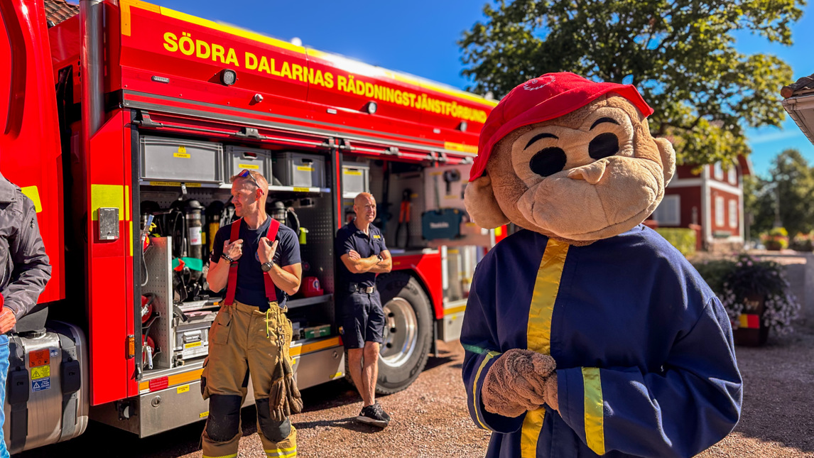 En maskot iklädd brandmanskläder står framför brandbil.