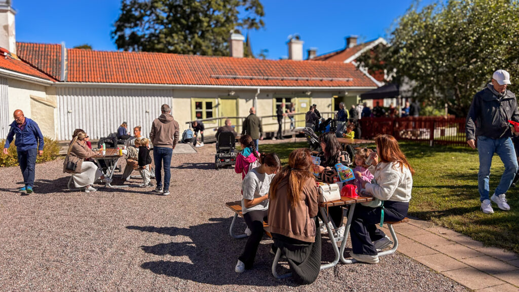 Många människor sitter på bänkar med bord utanför en äldre byggnad.