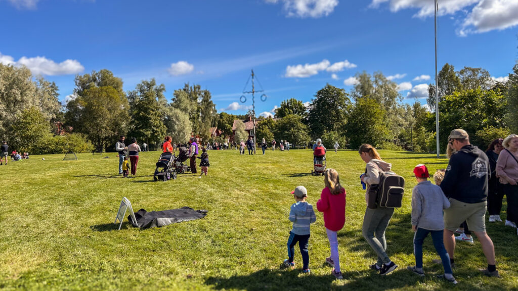 Många människor på en gräsyta runt en midsommarstång.